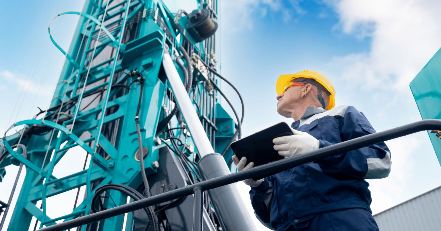 An oil rig operator looking up at machinery while holding a tablet